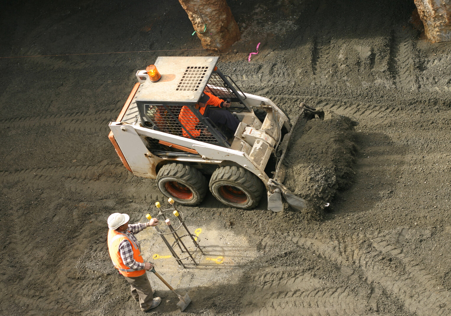 a bobcat shovels gravel while another workman ensures the site markings are kept clear.